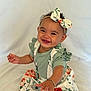 baby, barefoot, bow, bracelet, child, curly_hair, cute, floral_dress, green_clothing, happy, headband, indoors, infant, person, portrait, reaching_hand, sitting, smiling, white_background, young_child