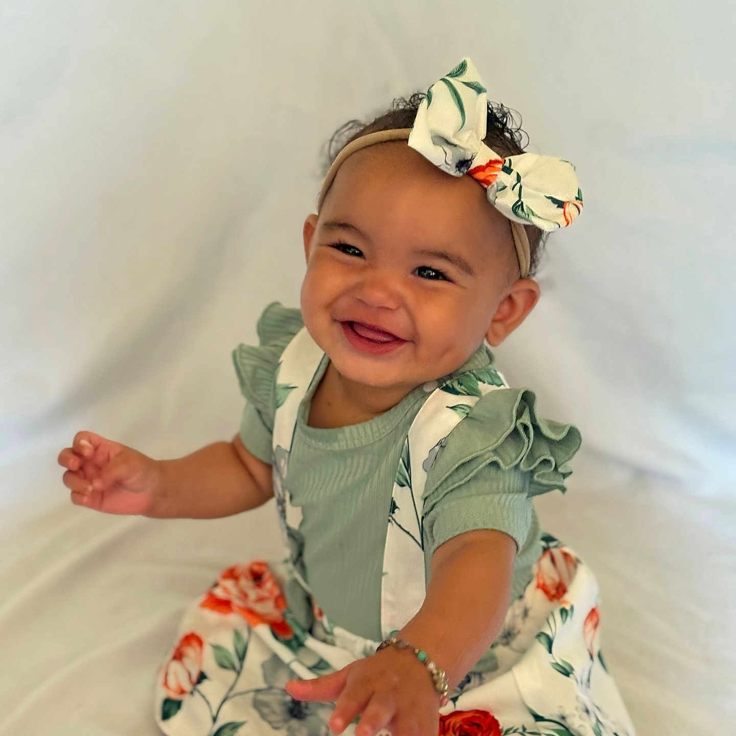 Nolani joined the competition — help win amazing prizes! baby, barefoot, bow, bracelet, child, curly_hair, cute, floral_dress, green_clothing, happy, headband, indoors, infant, person, portrait, reaching_hand, sitting, smiling, white_background, young_child