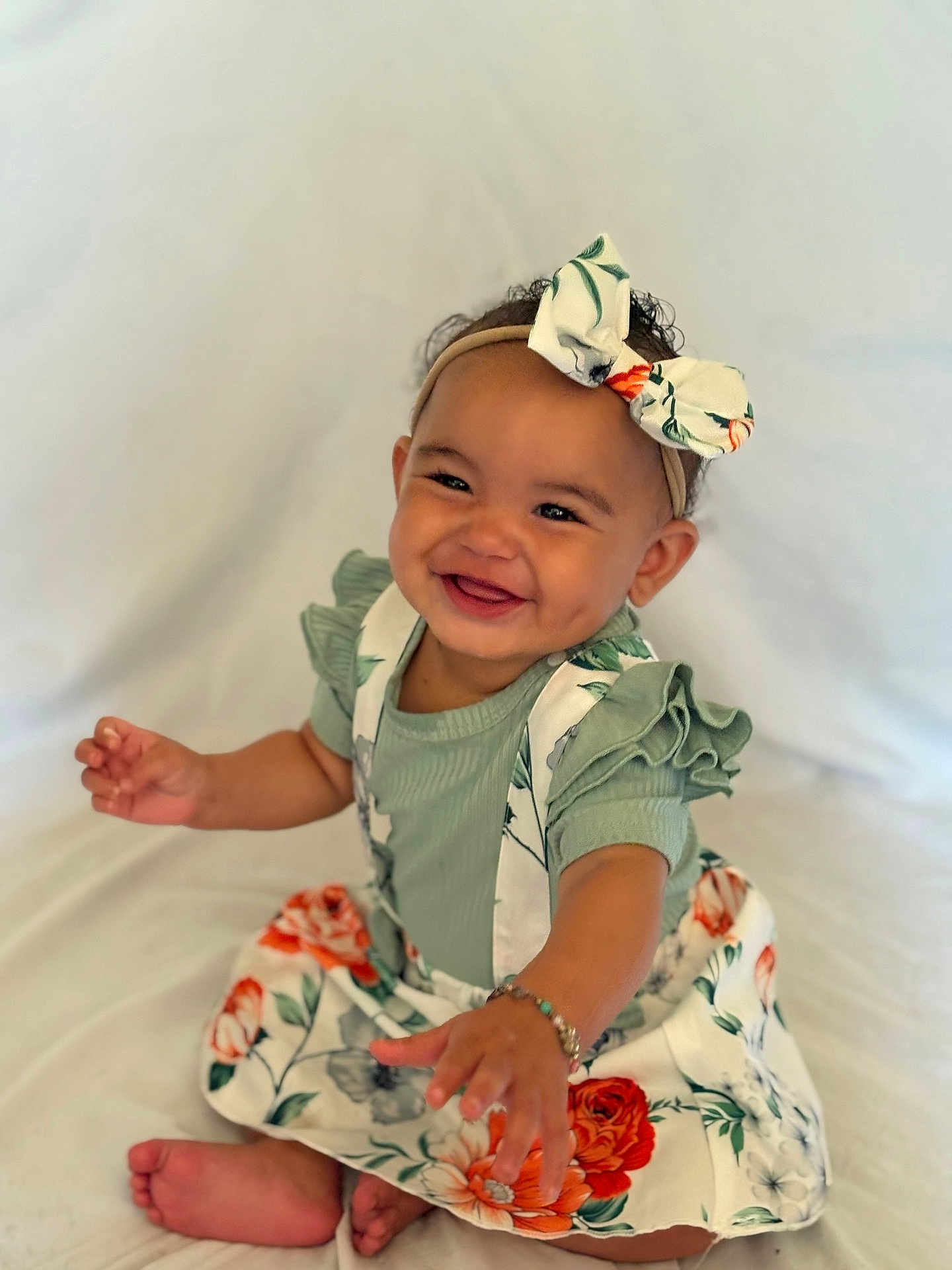 Nolani joined the competition — help win amazing prizes! baby, barefoot, bow, bracelet, child, curly_hair, cute, floral_dress, green_clothing, happy, headband, indoors, infant, person, portrait, reaching_hand, sitting, smiling, white_background, young_child