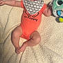 baby, bib, blanket, blue_eyes, child, cute, expression, face, foot, hand, headband, indoor, infant, lying_down, onesie, orange_clothing, person, plush_toy, smile, soft_toy
