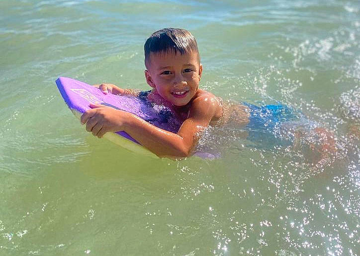 Ikaika is registered to the contest to win money with this photo: bathing, boardsport, facial_expression, fun, happy, joy, lake, leisure, ocean, outdoor_recreation, person, personal_protective_equipment, recreation, smile, sports, surfboard, surfing, surfing_equipment, toddler, water