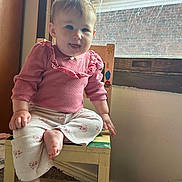 Elisheva is registered to the contest to win money with this photo: baby, child, blue_eyes, pink_clothing, floral_pants, wooden_chair, indoor, window, natural_light, playful_expression, tongue_out, carpet, wall, smiling, cute, person, sitting, home, young_child, casual