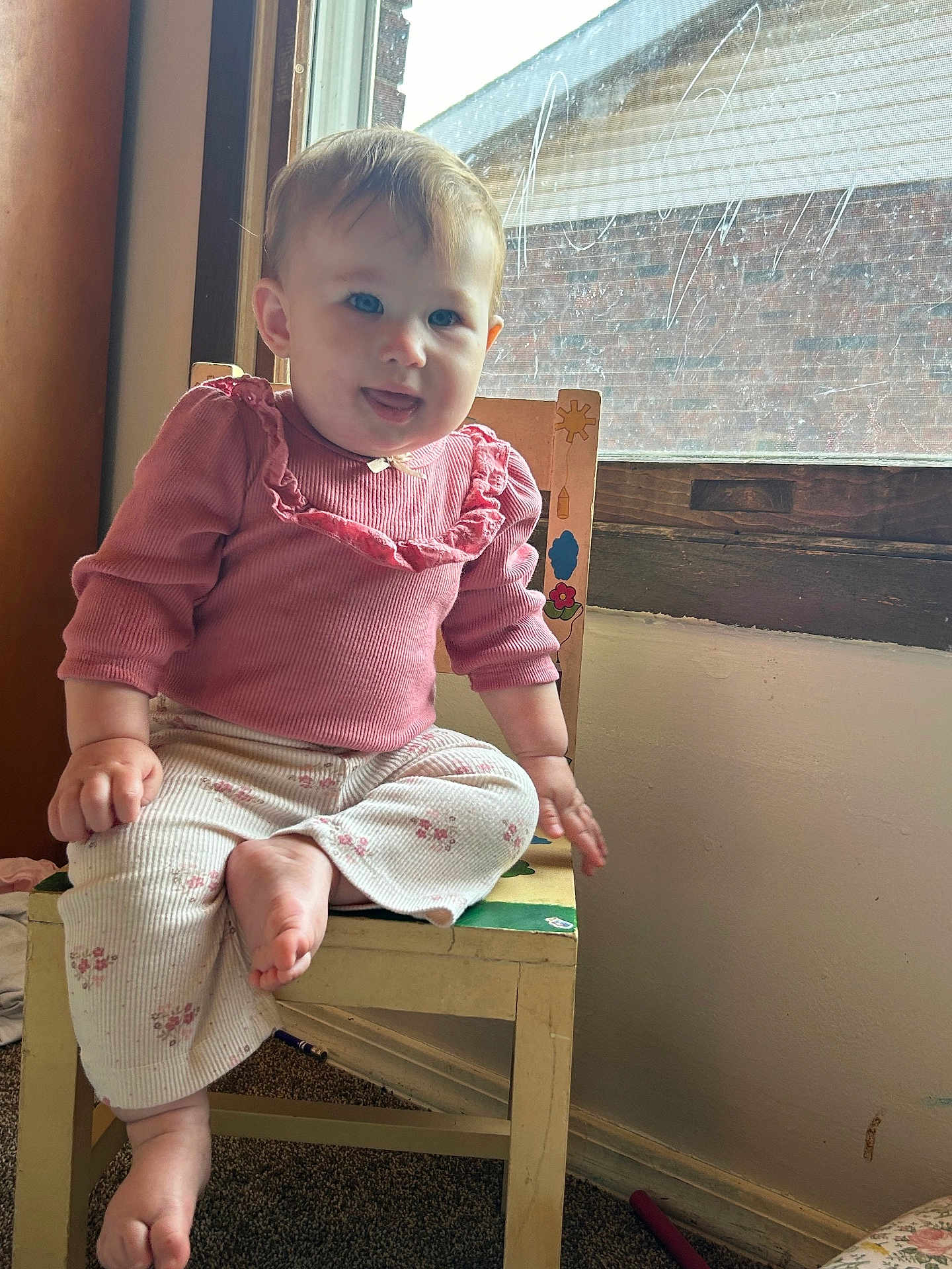 Elisheva is registered to the contest to win money with this photo: baby, child, blue_eyes, pink_clothing, floral_pants, wooden_chair, indoor, window, natural_light, playful_expression, tongue_out, carpet, wall, smiling, cute, person, sitting, home, young_child, casual