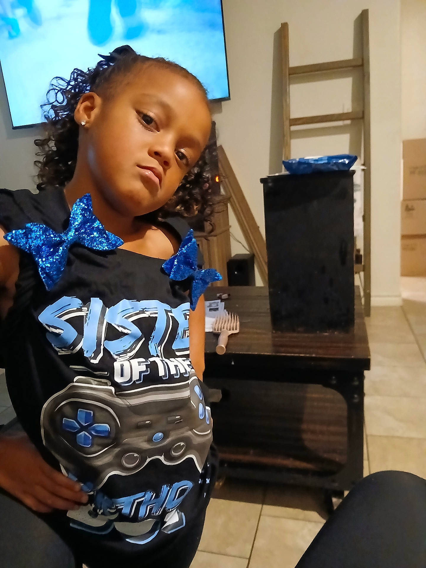 Zara joined the competition — help win amazing prizes! black_shirt, blue_bows, casual, child, comb, confident, curly_hair, earrings, floor_tiles, furniture, gaming_controller_graphic, girl, indoor, living_room, person, posing, sassy, table, television, wooden_ladder