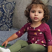 Naomi is registered to the contest to win money with this photo: bead_necklace, casual_clothing, child, couch, curly_hair, cushion, green_pants, home_interior, indoor, looking_at_camera, maroon_top, playing, portrait, relaxed, seated, sofa, textured_fabric, toddler, white_socks, young_child