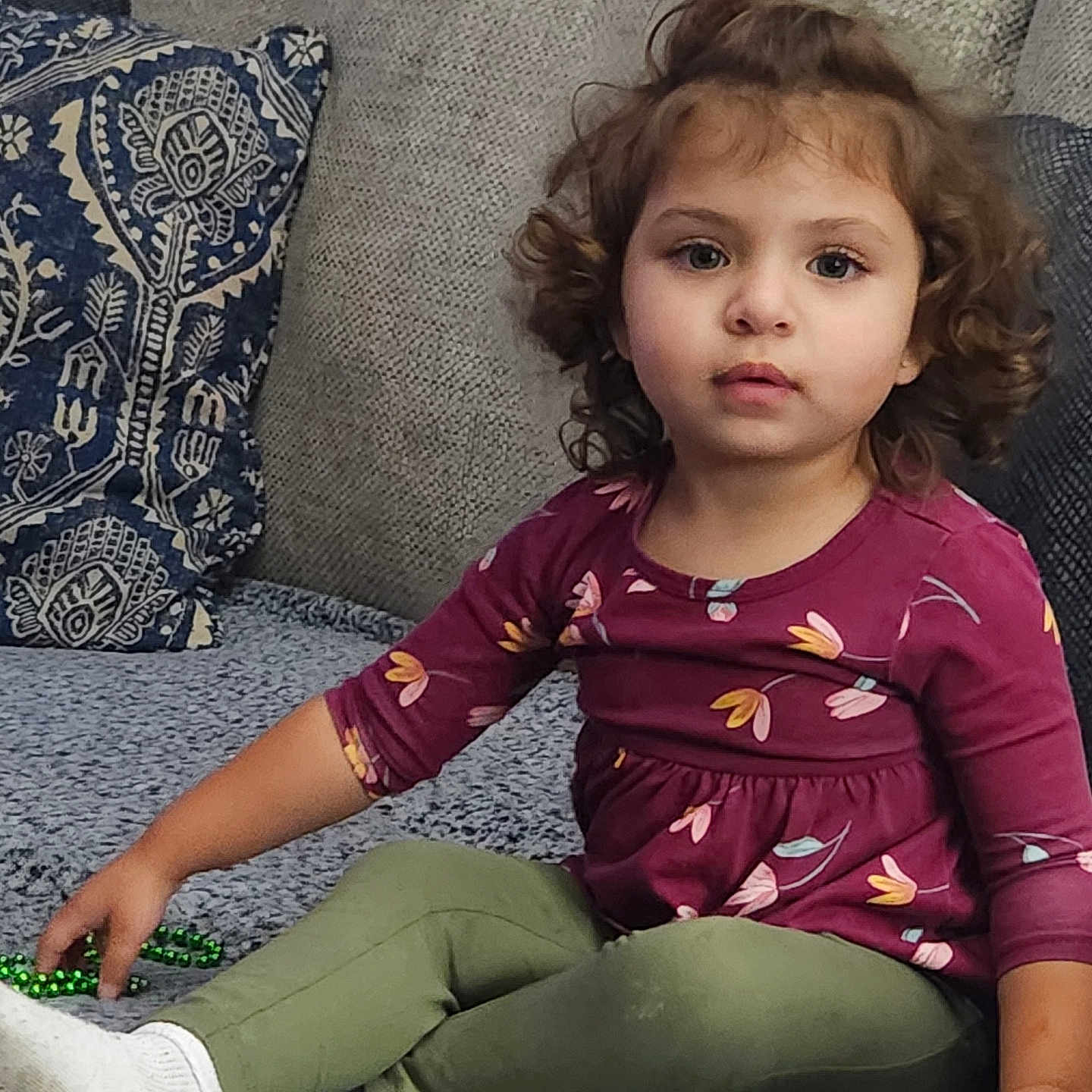 Naomi is registered to the contest to win money with this photo: bead_necklace, casual_clothing, child, couch, curly_hair, cushion, green_pants, home_interior, indoor, looking_at_camera, maroon_top, playing, portrait, relaxed, seated, sofa, textured_fabric, toddler, white_socks, young_child
