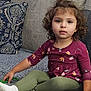 bead_necklace, casual_clothing, child, couch, curly_hair, cushion, green_pants, home_interior, indoor, looking_at_camera, maroon_top, playing, portrait, relaxed, seated, sofa, textured_fabric, toddler, white_socks, young_child