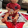 baby, toddler, child, sitting, red_clothing, striped_shirt, star_design, blanket, indoor, living_room, carpet, curly_hair, cute, holding_object, celebration, foot, face, expression, toy, decor