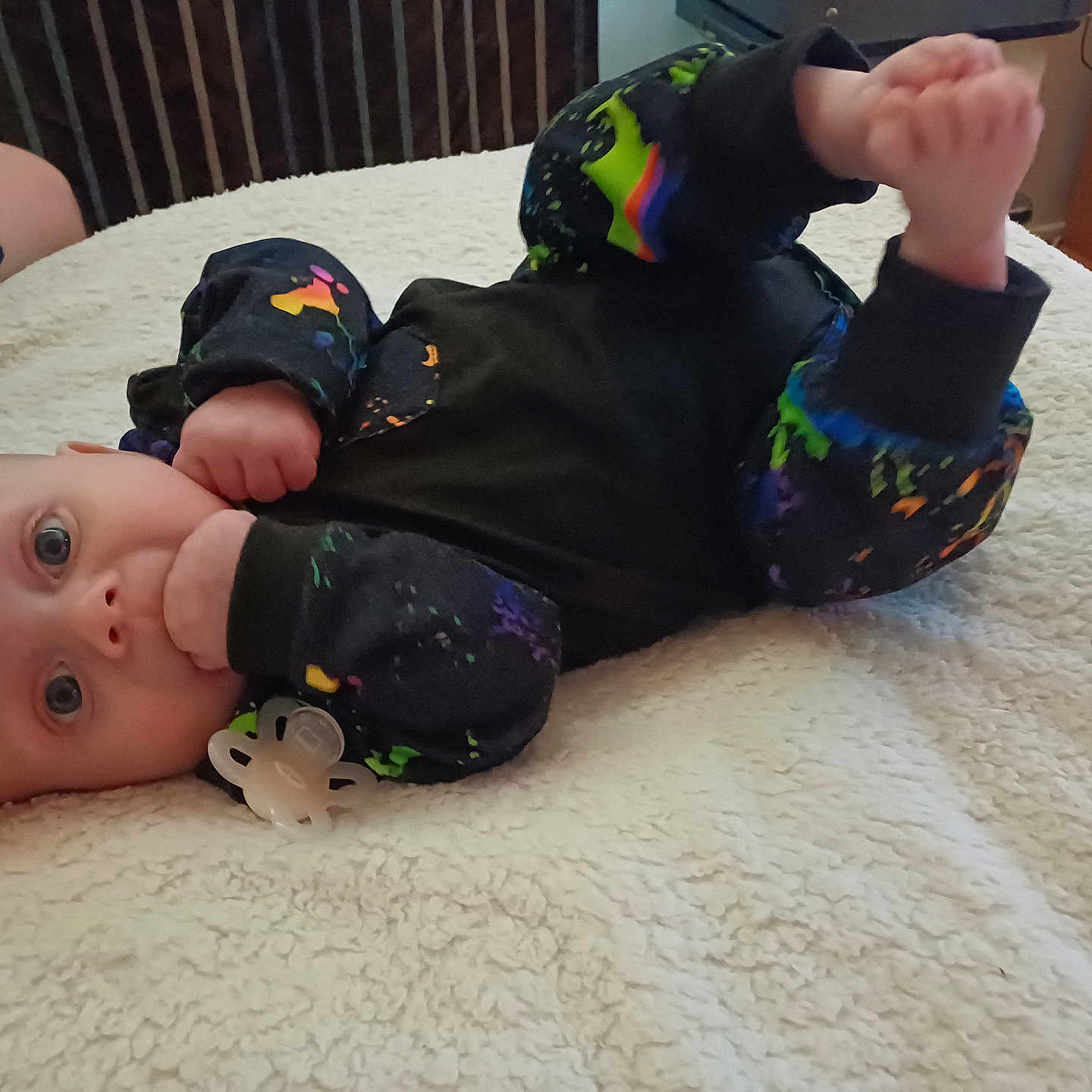 Jaydan is registered to the contest to win money with this photo: adult_hand, baby, bed, blanket, blue_eyes, casual, clothing, colorful, cute, face, fist, foot, hand, indoor, infant, lying_down, pacifier, person, phone, soft_texture