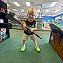 black_shirt, blond_hair, casual_clothing, ceiling_lights, child, collectibles, floor, green_carpet, happy, indoor, neon_shoes, person, playful, retail, shelves, shorts, smiling, standing, store, toy_hammer