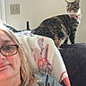 woman, glasses, cat, tabby_cat, indoor, couch, blanket, clock, wall, relaxed, home, furniture, portrait, seated, light_switch, casual, pet, companion, comfort, smile