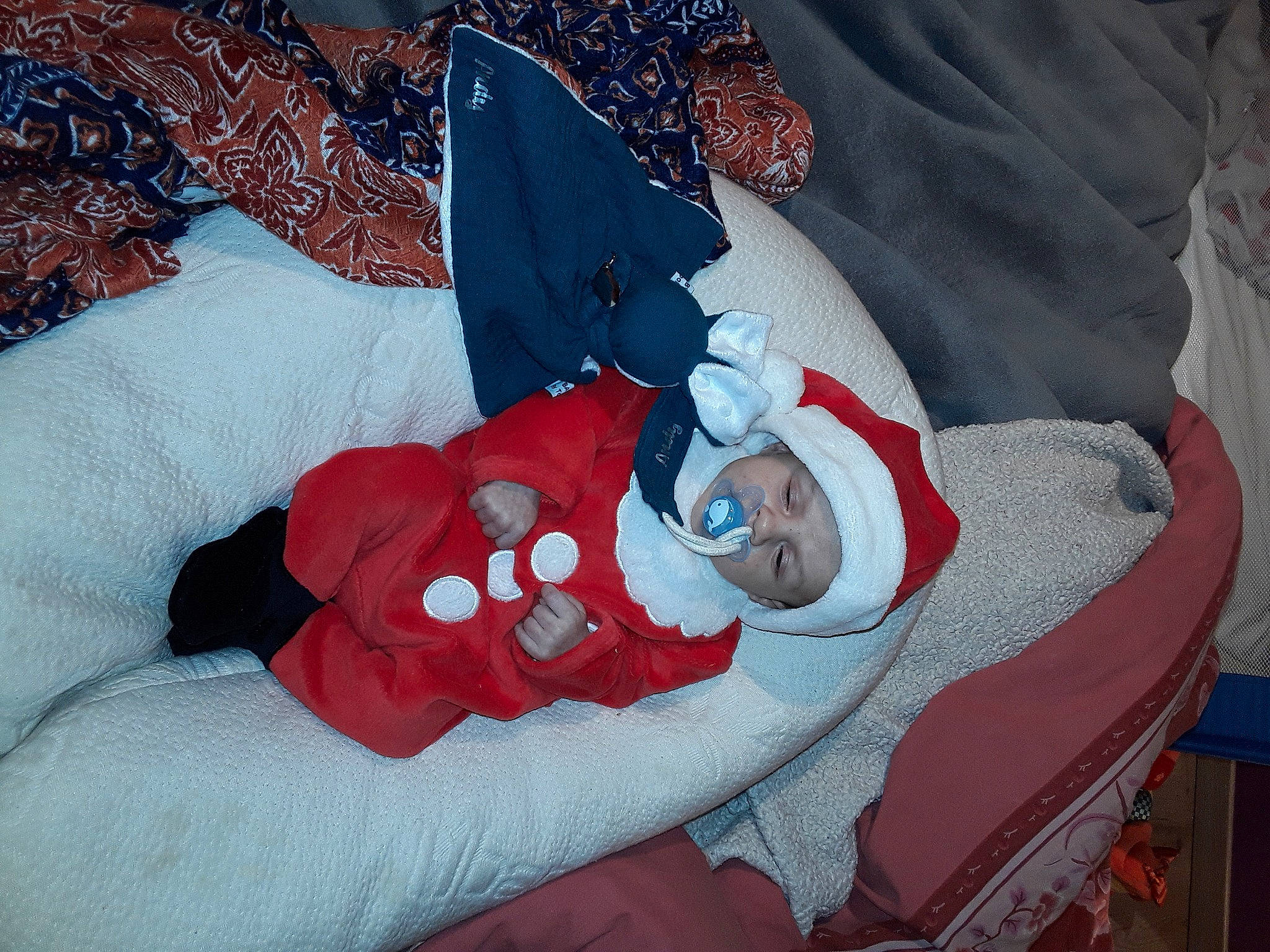 Andy participe au concours pour gagner de l'argent avec cette photo : baby, baby_products, baby_sleeping, baby_toddler_clothing, carmine, child, comfort, costume, event, fictional_character, hat, head, headwear, human_body, lap, linens, pattern, person, room, sleeve