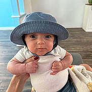 Cash joined the competition — help win amazing prizes! baby, child, hat, blue_hat, white_shirt, blanket, indoor, hand, red_nail_polish, curious, wide_eyes, plump, person, infant, holding, couch, wooden_floor, face, portrait, cute