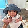 baby, child, hat, blue_hat, white_shirt, blanket, indoor, hand, red_nail_polish, curious, wide_eyes, plump, person, infant, holding, couch, wooden_floor, face, portrait, cute