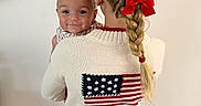 Cash joined the competition — help win amazing prizes! baby, child, braid, red_bow, sweater, american_flag, blue_shorts, smile, face, person, hair, indoor, holding, cute, portrait, young_child, white_wall, casual_clothing, standing, back_view