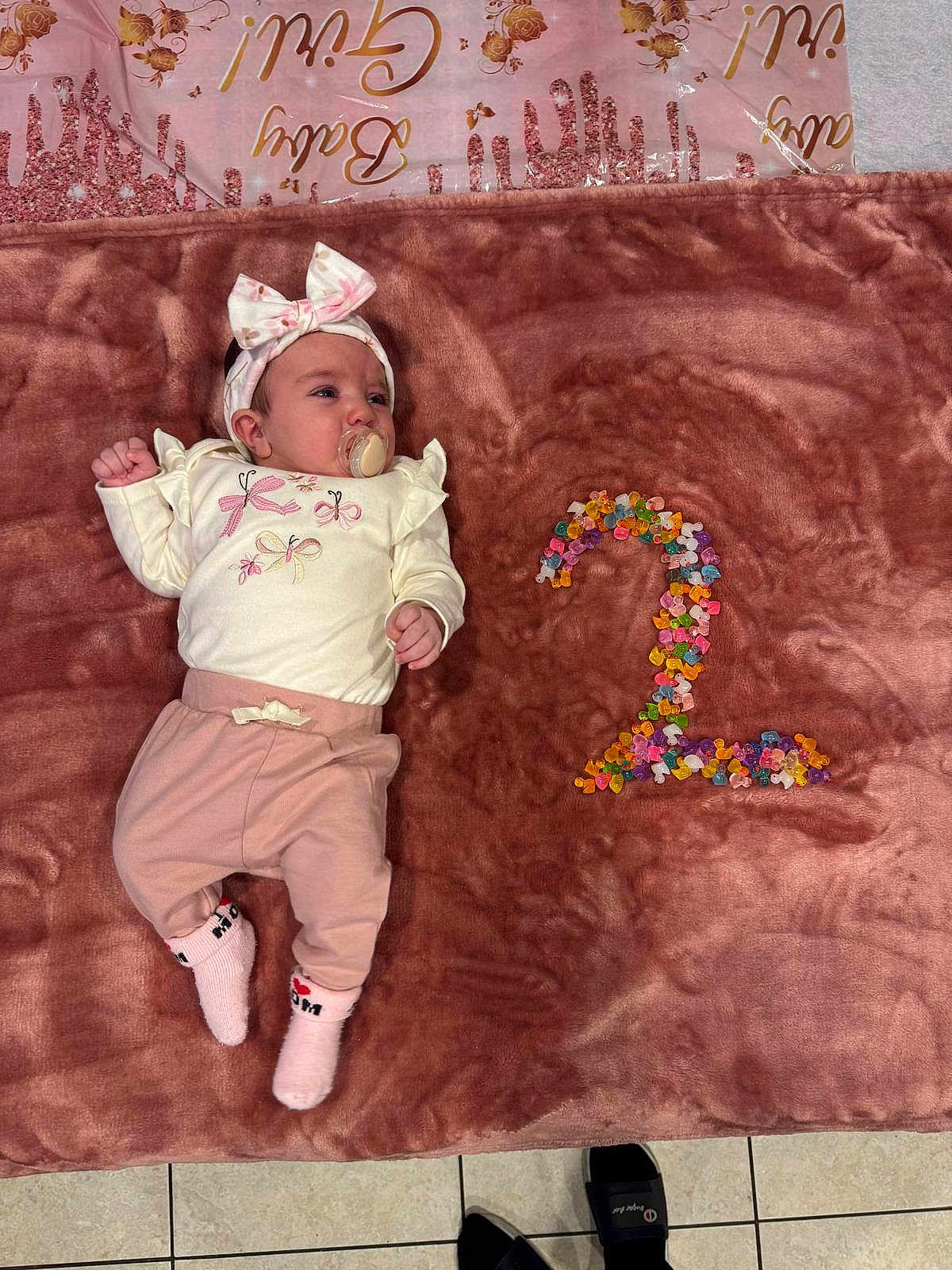 Lia Vana joined the competition — help win amazing prizes! baby, girl, pacifier, headband, blanket, pink_clothing, white_top, beads, number_two, floor_tiles, indoor, cute, child, infant, lying_down, soft_texture, colorful, decor, small_person, socks