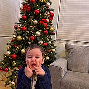 Hovhannes Garbis is registered to the contest to win money with this photo: child, christmas_tree, holiday, decorations, red_ornaments, gold_ornaments, lights, living_room, couch, window_blinds, floor_tiles, wrapped_gift, silly_face, blue_jacket, playful, person, indoor, smile, festive, home