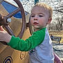 Jonathan is registered to the contest to win money with this photo: toddler, child, playground, steering_wheel, outdoor, daylight, green_sleeves, blue_eyes, person, toy, play_equipment, park, trees, bench, clothing, hand, face, curious, casual, young_child