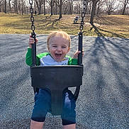 Jonathan is registered to the contest to win money with this photo: child, toddler, swing, playground, outdoor, smiling, happy, sunlight, trees, shadow, grass, park, casual_clothing, blue_pants, green_shirt, white_socks, shoes, daylight, chain, seat