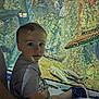 child, toddler, aquarium, fish, glass, underwater, rocks, plaid_shorts, gray_shirt, sneakers, informational_display, sea_life, water, sitting, curious, indoor, exhibit, person, blue_shoes, education