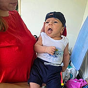 Mael a rejoint le concours — aidez-le/la à gagner de superbes lots ! baby, child, woman, holding, hat, cap, white_shirt, shorts, socks, indoor, laundry, snack, cheetos, red_shirt, face, expression, person, clothing, home, casual