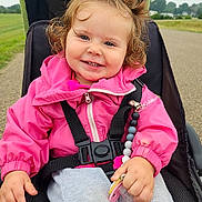 Harlee is registered to the contest to win money with this photo: child, clothing, curly_hair, cute, daytime, face, grass, happy, nature, outdoor, pacifier, person, pink_jacket, portrait, road, seatbelt, smiling, stroller, toddler, toy