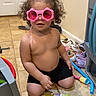 toddler, child, pink_sunglasses, curly_hair, book, tile_floor, indoor, shoes, toy, kneeling, black_shorts, playtime, cute, young_child, home, colorful, floor, reading, fun, casual