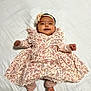 baby, infant, smiling, dress, floral_pattern, headband, lying_down, white_blanket, happy, cute, child, portrait, indoors, soft_lighting, small_hands, small_feet, fabric, patterned_clothing, adorable, ruffles
