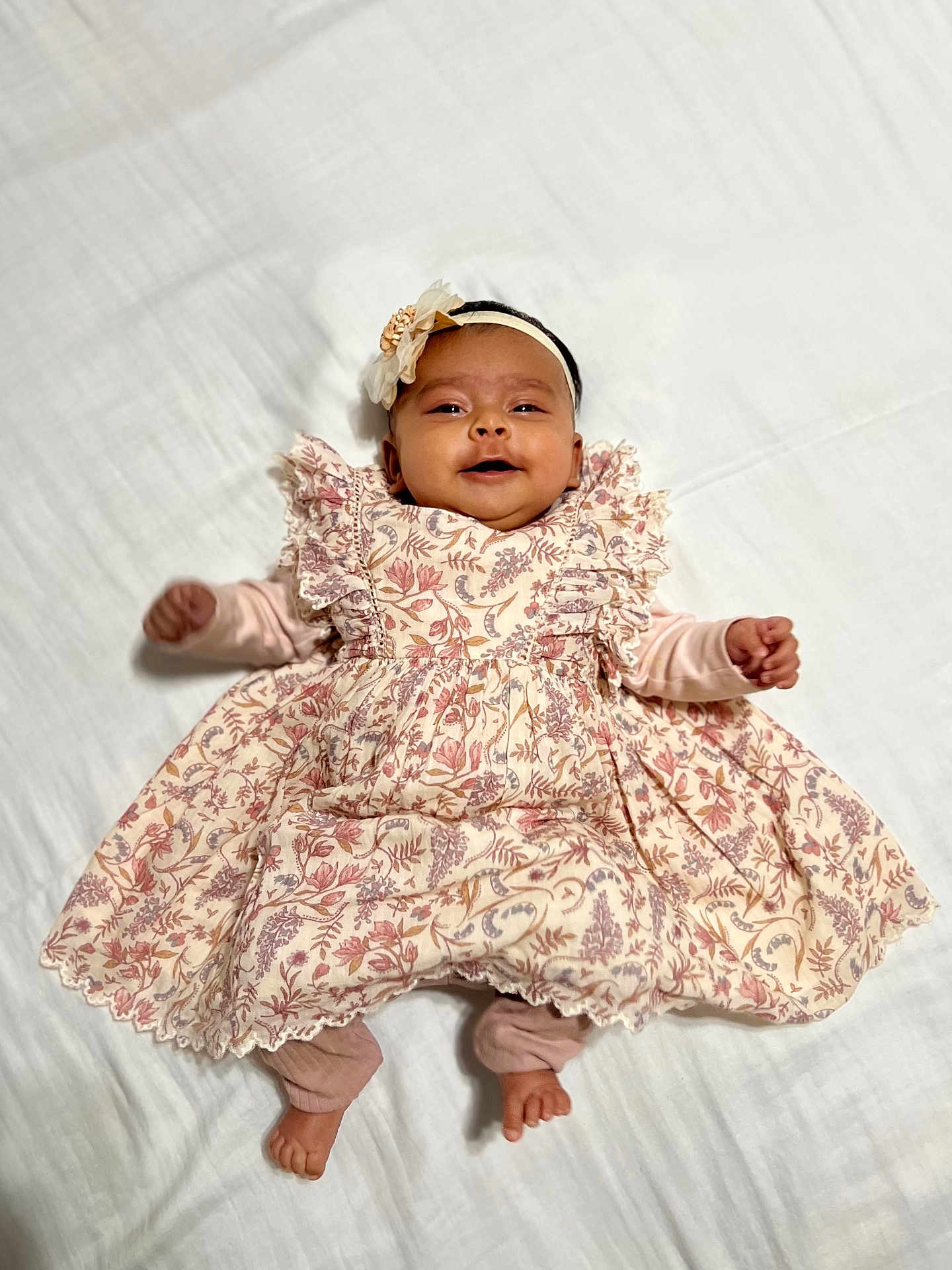 Evana joined the competition — help win amazing prizes! baby, infant, smiling, dress, floral_pattern, headband, lying_down, white_blanket, happy, cute, child, portrait, indoors, soft_lighting, small_hands, small_feet, fabric, patterned_clothing, adorable, ruffles