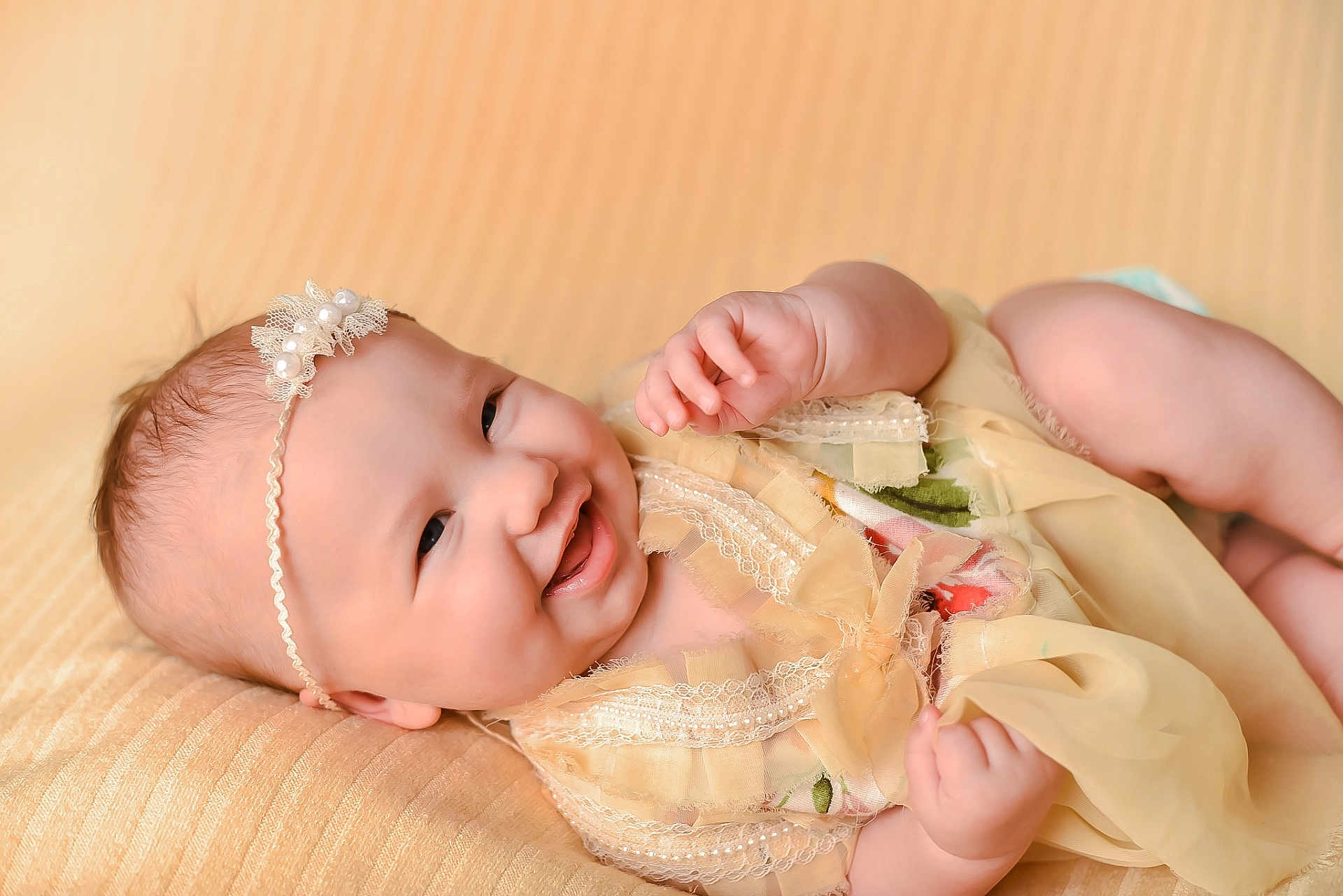 Rora is registered to the contest to win money with this photo: baby, smiling, infant, headband, lace, yellow_dress, cute, portrait, soft_texture, happy, child, newborn, skin, closeup, lying_down, fabric, pearl, adorable, expression, indoors