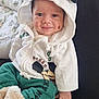 baby, child, hoodie, smile, cute, green_pants, white_clothing, face, person, indoor, blanket, sitting, happy, clothing, infant, cozy, lap, soft_toy, hood, casual