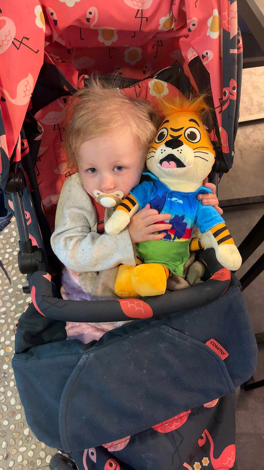 Kyra Griffin is registered to the contest to win money with this photo: child, stroller, pacifier, plush_toy, tiger_toy, flamingo_pattern, clothing, indoor, cute, baby, toy, fabric, seat, comfort, person, blond_hair, hand, face, sitting, expression