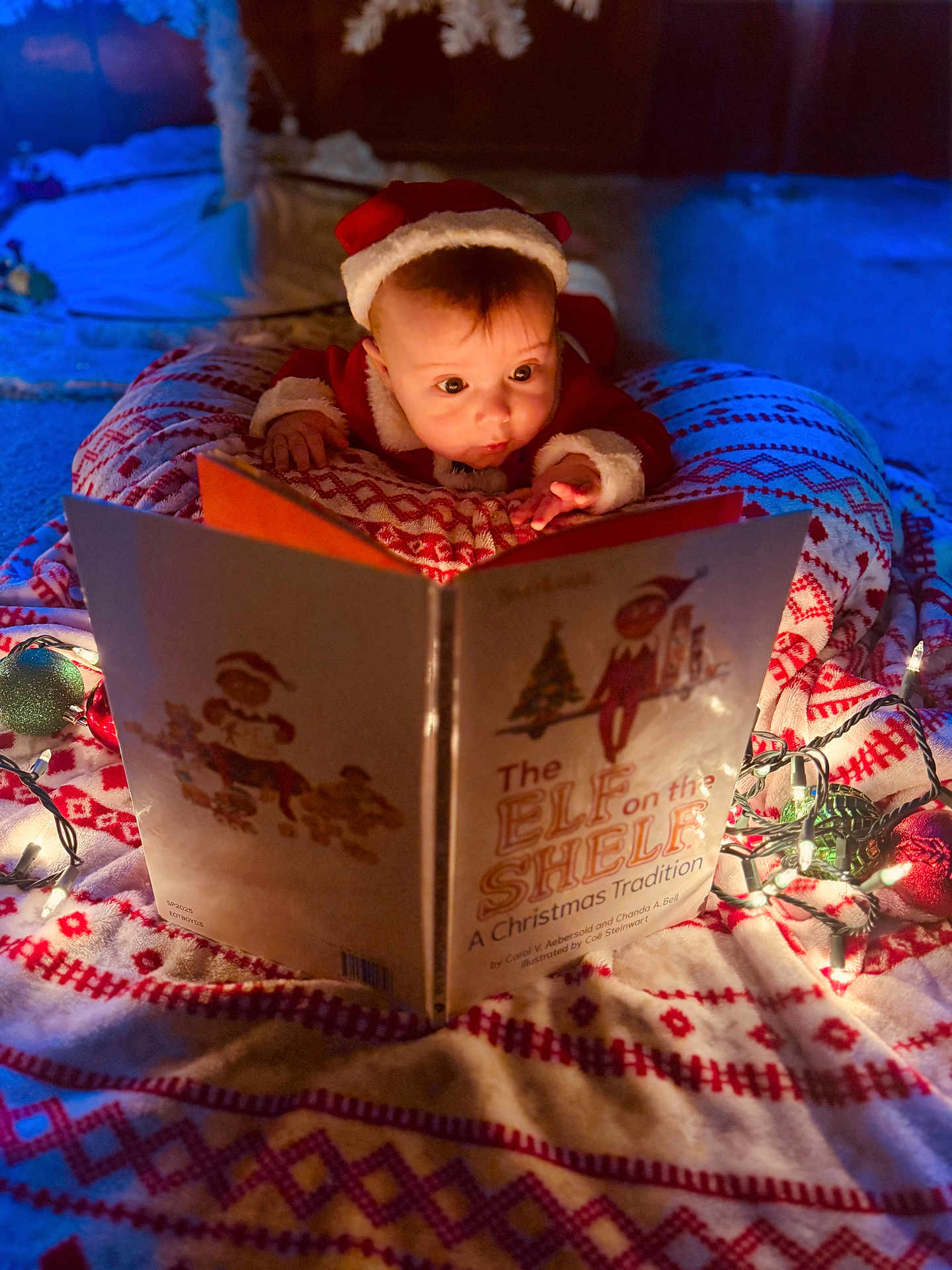 Ian joined the competition — help win amazing prizes! baby, child, christmas, holiday, santa_hat, book, reading, blanket, lights, decorations, festive, indoors, cute, face, portrait, infant, warm_lighting, cozy, holiday_spirit, toddler