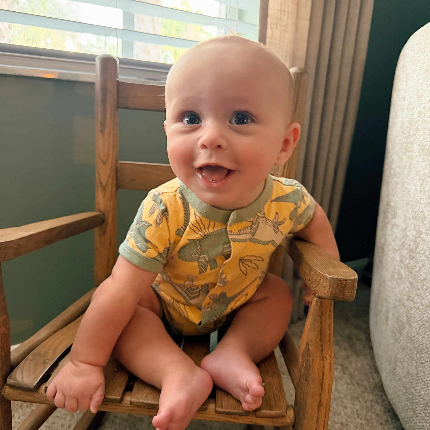 Beau joined the competition — help win amazing prizes! baby, child, smile, dinosaur_print, onesie, wooden_chair, rocking_chair, indoor, window_blinds, natural_light, carpet, curtains, happy, cute, infant, seated, person, furniture, home, portrait
