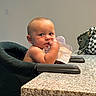 baby, high_chair, countertop, paper, bag, checkered_pattern, table, indoor, child, face, skin, person, curious, sitting, hand, arm, head, light, wall, furniture