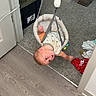 baby, doorway_jumper, infant, carpet, floor, doorway, clothing, indoor, child, play, suspended, curious, expression, home, wood_floor, plastic, fabric, toy, baby_gear, childcare