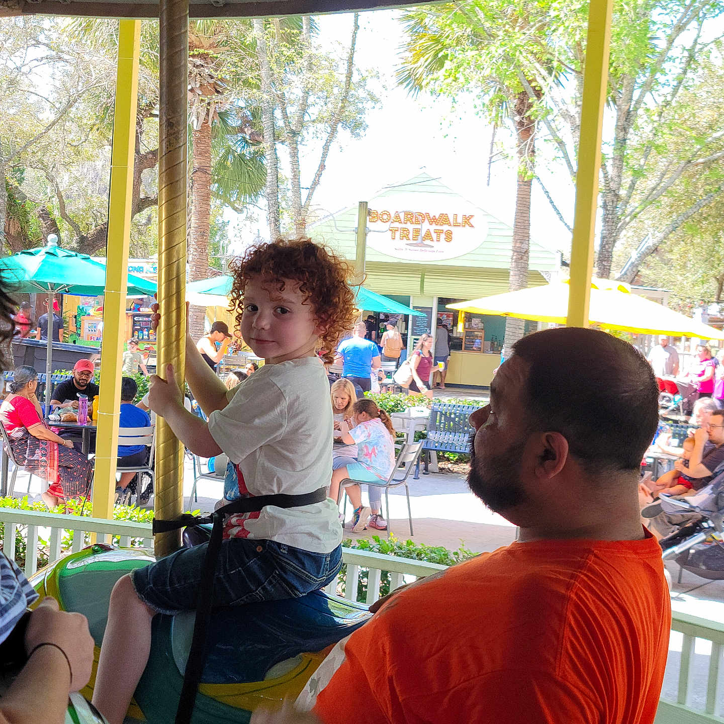 Lucas joined the competition — help win amazing prizes! amusement_park, boardwalk_treats, carousel, casual_clothing, child, curly_hair, daytime, fence, fun, horse, man, orange_shirt, outdoor, park, people, pole, seating, sign, trees, umbrella