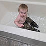 bathroom, bathtub, camo_pants, candid, casual, child, curious_expression, floor_tiles, home, indoor, person, plumbing, real_photo, short_hair, sitting, skin, toddler, water, wet_clothing, young_child