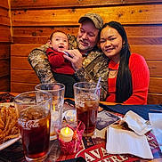 Brentley is registered to the contest to win money with this photo: baby, camouflage, candle, coca_cola, drink, family, fried_food, glasses, hat, indoor, man, napkins, plate, portrait, red_sweater, restaurant, smiling, table, woman, wooden_wall