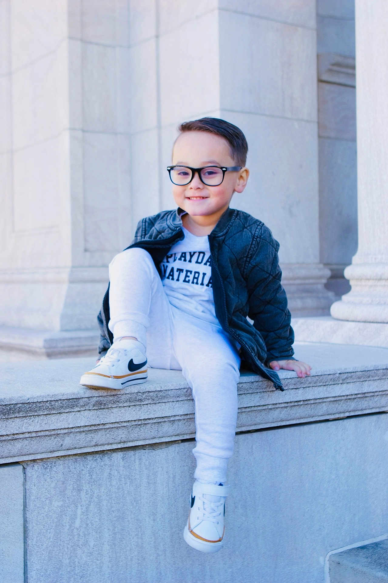 Asher is registered to the contest to win money with this photo: child, boy, smile, glasses, jacket, sneakers, sitting, stone, ledges, building, architecture, outdoor, casual, happy, person, fashion, young, portrait, daytime, urban
