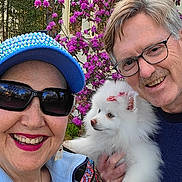 Betty Lou joined the competition — help win amazing prizes! adult, smiling, outdoor, dog, white_dog, fluffy_dog, glasses, hat, jacket, flower, pink_flower, porch, grass, happy, pet, holding, portrait, nature, people, fun