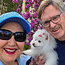 adult, smiling, outdoor, dog, white_dog, fluffy_dog, glasses, hat, jacket, flower, pink_flower, porch, grass, happy, pet, holding, portrait, nature, people, fun