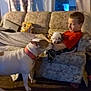 boy, puppy, dog, couch, blanket, carpet, indoor, curtains, window, food_bowl, child, pet, animal, furniture, red_shirt, sniffing, holding, cozy, home, playing
