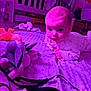 baby, infant, blanket, plush_toy, stuffed_animal, purple_light, crib, carpet, clothing, face, person, indoors, child, toy, soft_texture, wide_eyes, floor, playtime, cute, curious
