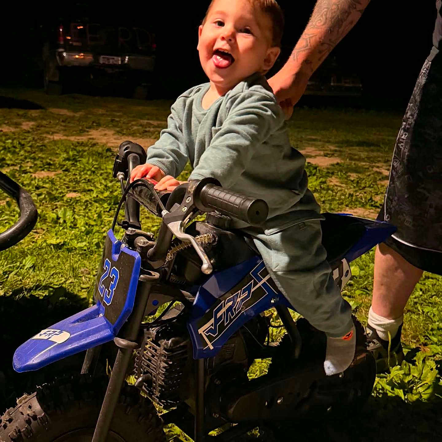 Wyatt is registered to the contest to win money with this photo: child, toddler, dirt_bike, toy_vehicle, grass, night, adult_hand, tattoo, playful, outdoor, happy, motorbike, small_vehicle, person, fun, clothing, footwear, dark_background, illuminated, tongue_out