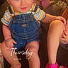 child, baby, toddler, denim_overalls, pink_shoes, footwear, indoor, couch, person, smile, happy, face, legs, sitting, clothing, timestamp, text_overlay, hair, casual, warm_lighting