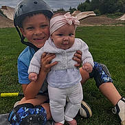 Maya joined the competition — help win amazing prizes! child, baby, boy, helmet, protective_gear, knee_pads, grass, outdoor, smile, headband, sweater, playground, skate_park, happy, siblings, casual_clothing, fun, nature, daytime, park