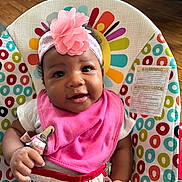 Alana is registered to the contest to win money with this photo: baby, smiling, headband, pink_bib, baby_seat, colorful_pattern, elephant_design, hand, clip, infant, face, cute, indoors, wood_floor, happy, child, portrait, toddler, clothing, seat