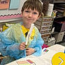 Hunter joined the competition — help win amazing prizes! child, classroom, school, student, smiling, portrait, blue_smock, yellow_shirt, brown_hair, table, worksheet, paper, pencil, marker, art_supplies, learning, education, classroom_decor, shelves, indoor