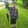 child, boy, fishing, fish, angler, outdoor, grass, lawn, trees, nature, smiling, portrait, suburban, sidewalk, houses, stop_sign, jacket, camouflage, shorts, digital_scale